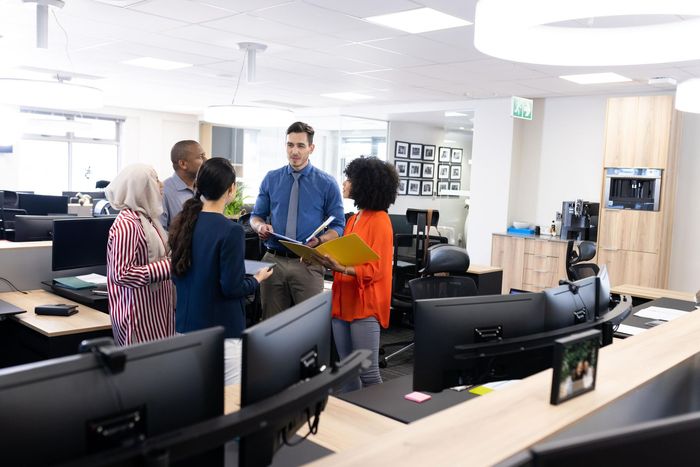 Group of people in an office discussing documents; bright, open space with computers.