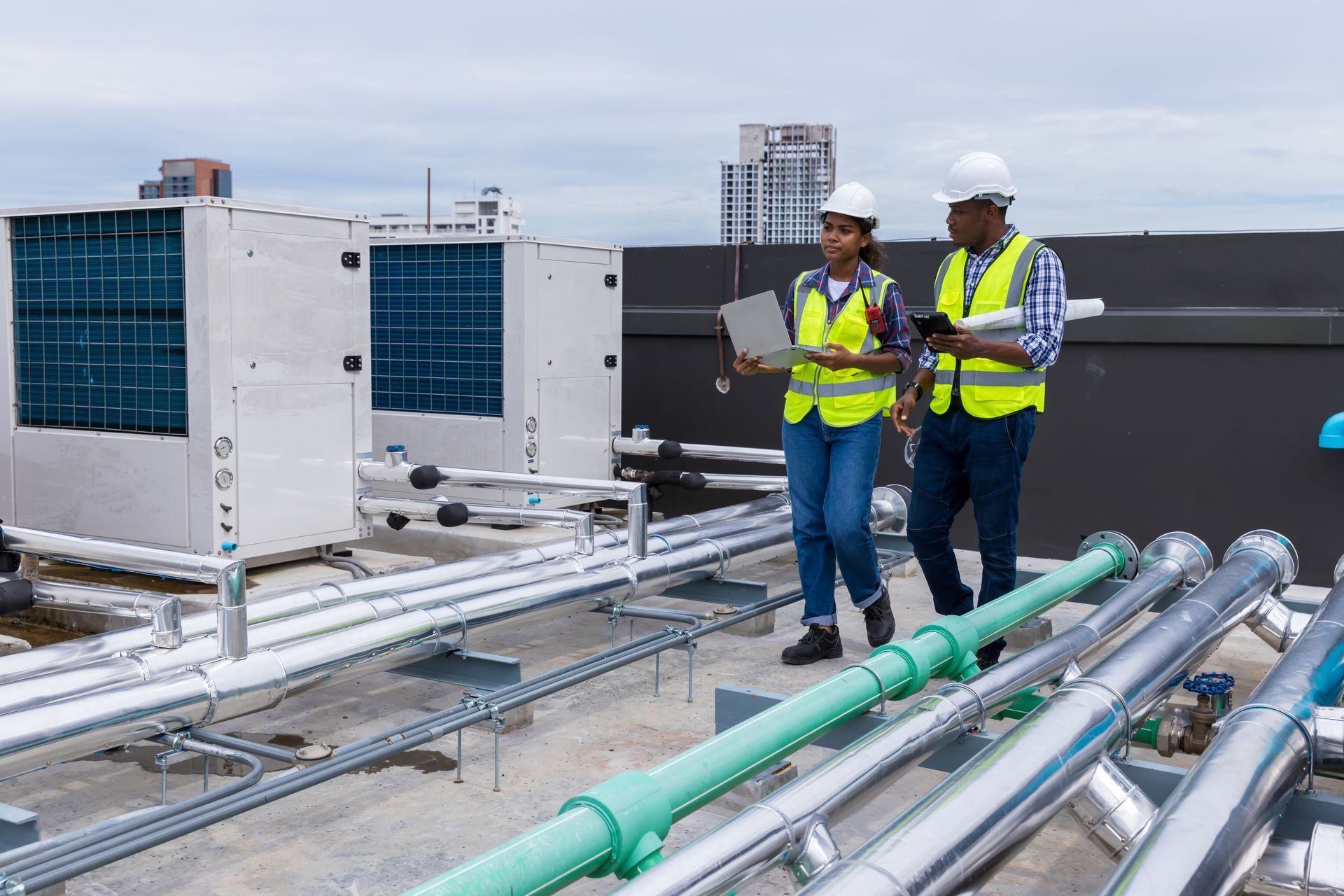 Two people in safety vests and hard hats inspect HVAC equipment on a rooftop.