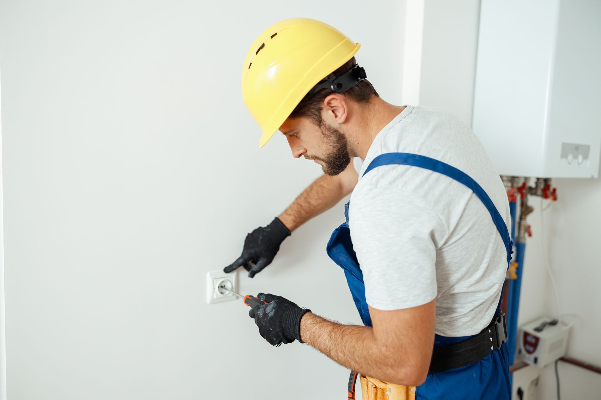 Electrician in a yellow hard hat working on an electrical outlet on a white wall.