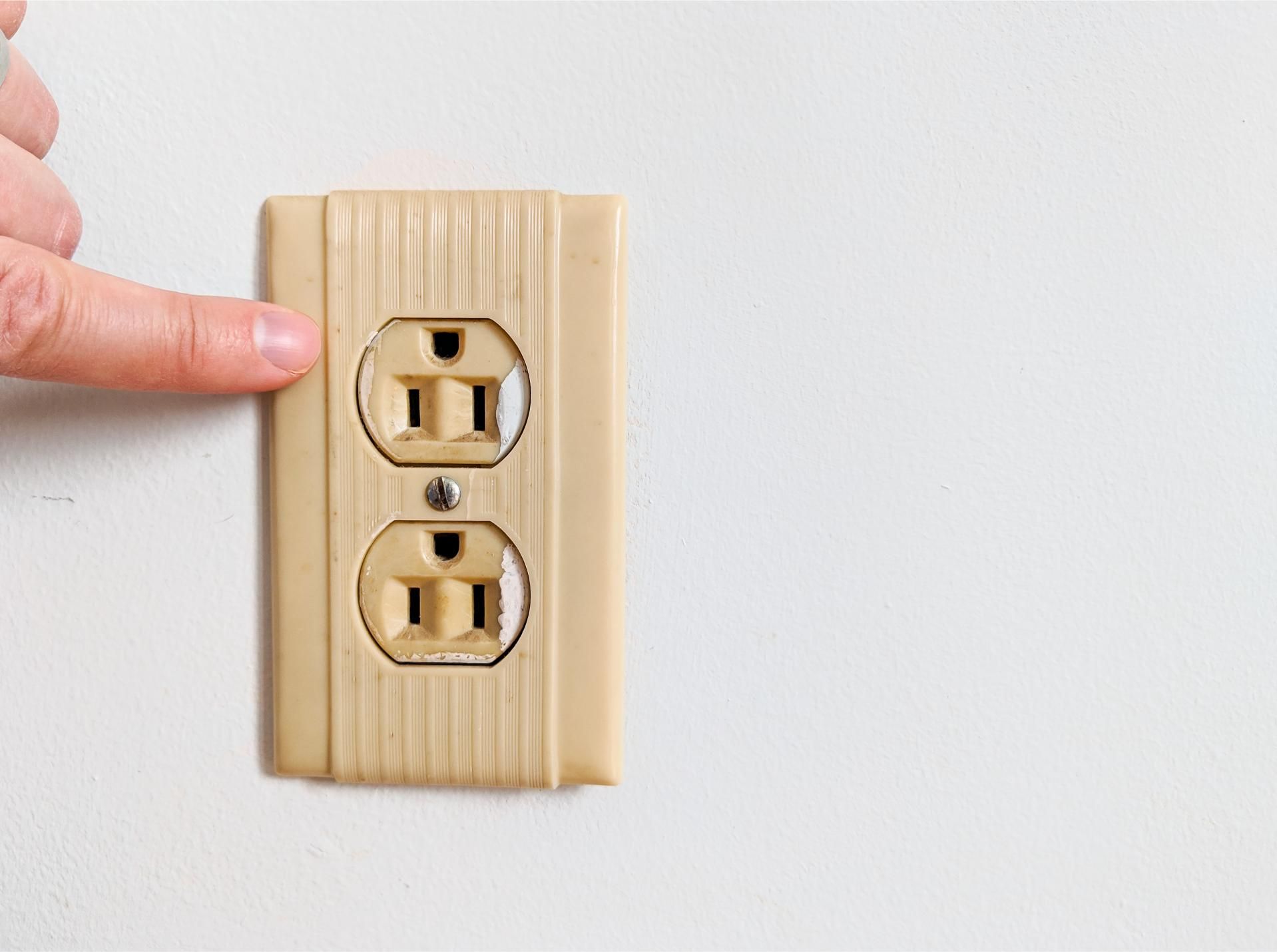 Finger touching a beige electrical outlet on a white wall.
