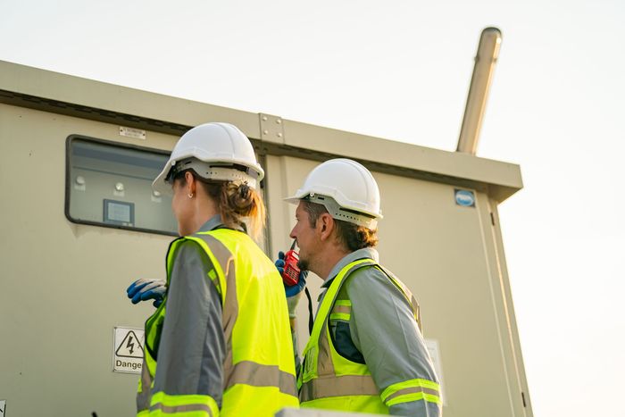 Two workers in safety vests and hard hats inspecting electrical equipment outdoors.