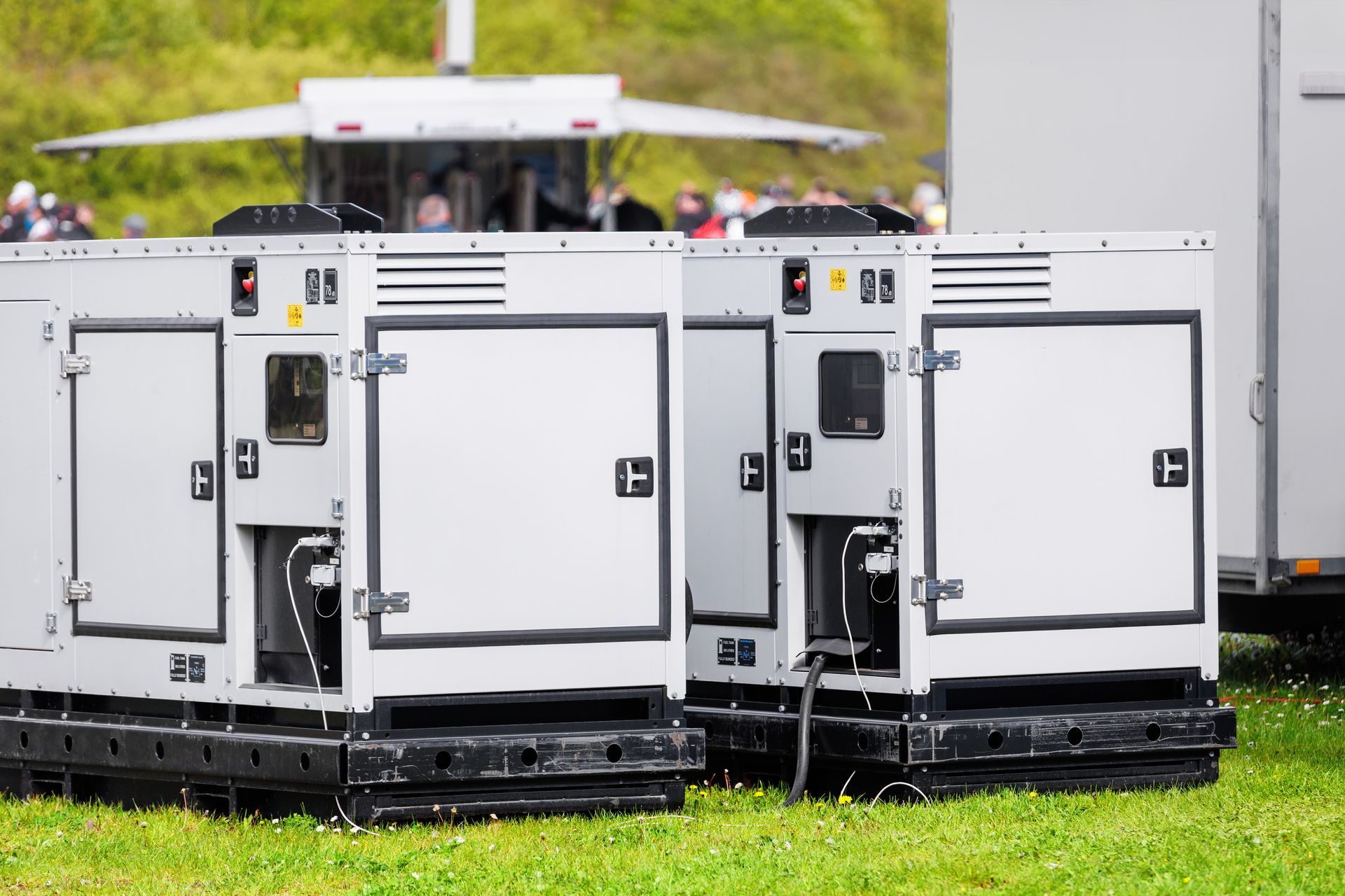 White portable generator units on wheeled bases outdoors at an event, with crowd and equipment in the background