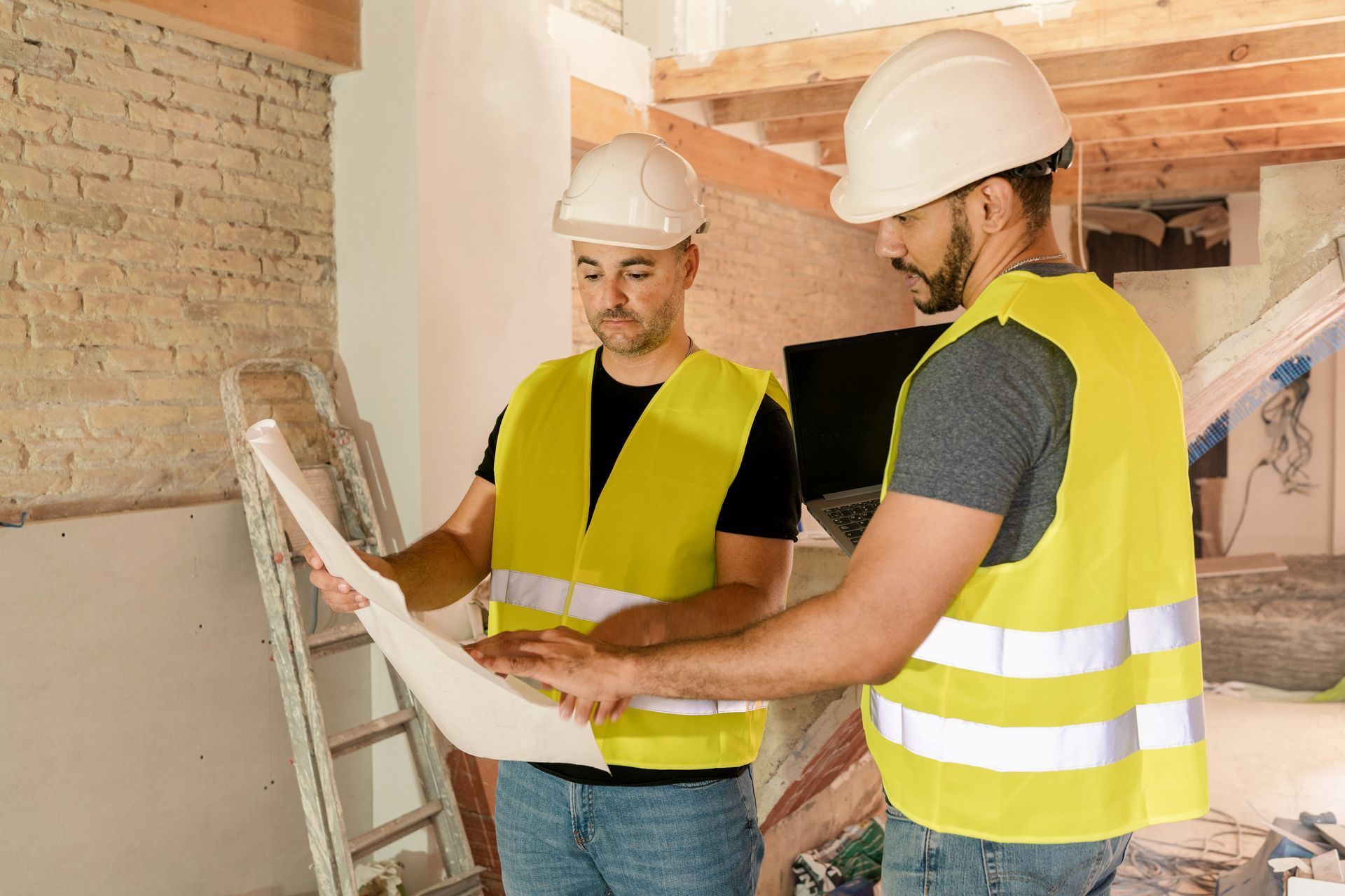 Two construction workers in vests and hard hats reviewing blueprints at a construction site.