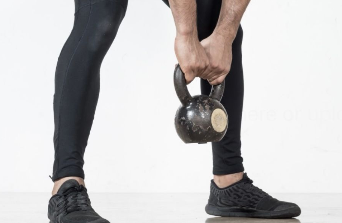 A person wearing black leggings and sneakers holds a kettlebell with both hands while in a bent-over position.