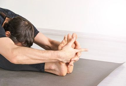A person in a seated forward fold yoga pose, holding their feet with both hands on a gray mat.