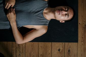 A person in a gray tank top lying in Savasana on a dark yoga mat over a wooden floor.