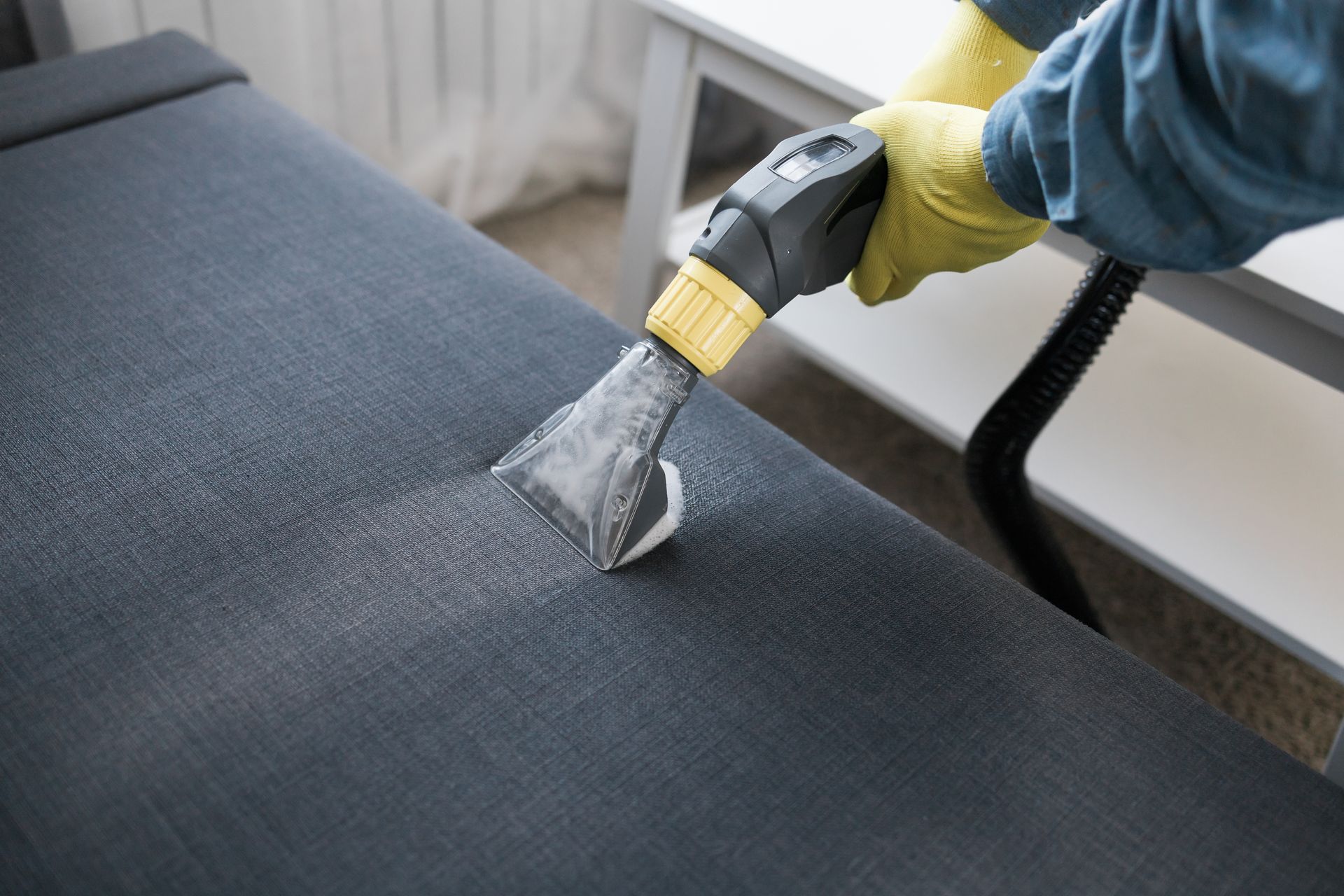 A person wearing yellow gloves steam cleans a gray upholstered surface.