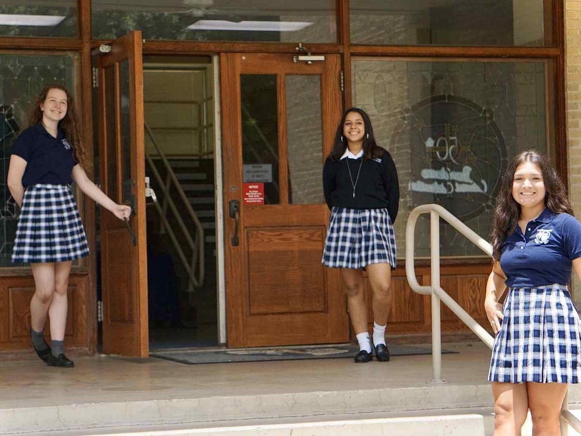 Three girls in plaid skirts stand in front of a building