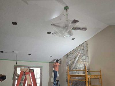 People painting a ceiling, covering a ceiling fan and a stone wall with plastic sheeting.