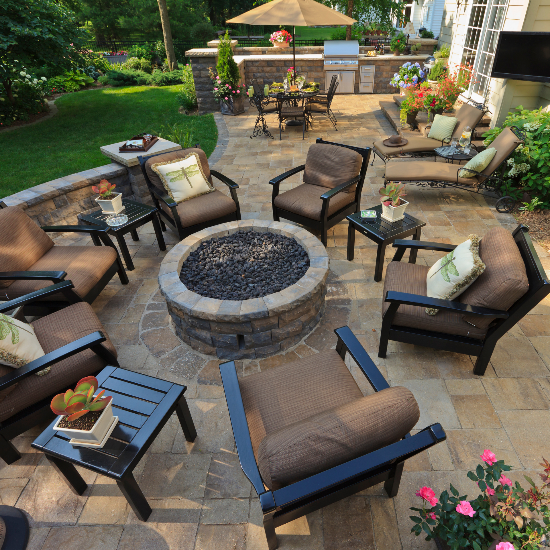 Patio with fire pit, seating, grill, and dining area. Stone paving, brown furniture, green lawn, sunny day.
