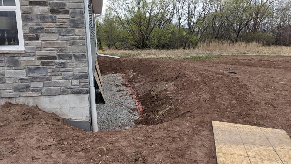 A house foundation being prepared for gravel, with a trench, dirt piles, and a piece of wood.
