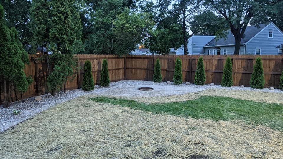 A backyard with a brown wooden fence, small trees, grass, and gravel landscaping.