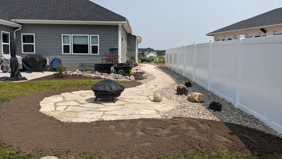 Backyard with stone patio, fire pit, and pathway along a white fence.