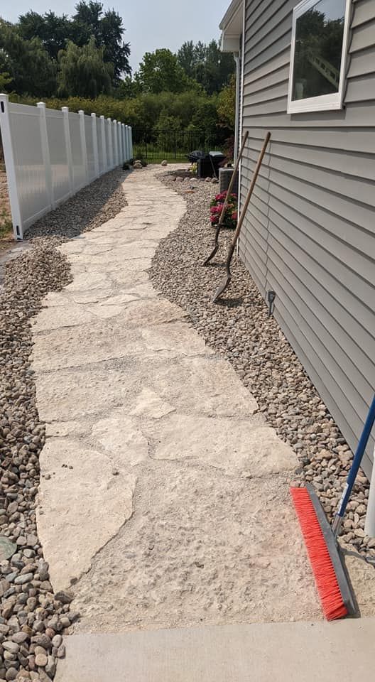 Stone path with rock border next to a house with siding and a white fence on a sunny day.