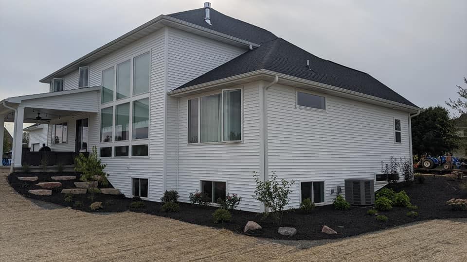 White two-story house with dark roof and trim. Landscaping with black mulch borders the house.