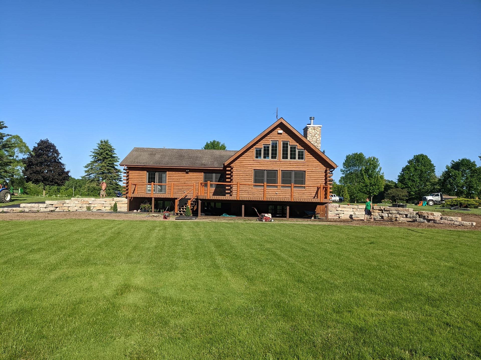 Wooden cabin with deck, chimney, and large windows on a grassy lawn under a clear blue sky.
