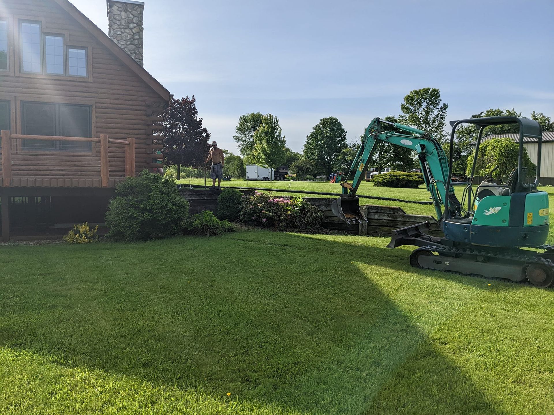 Small excavator digging in a grassy yard next to a log cabin, under a sunny sky.