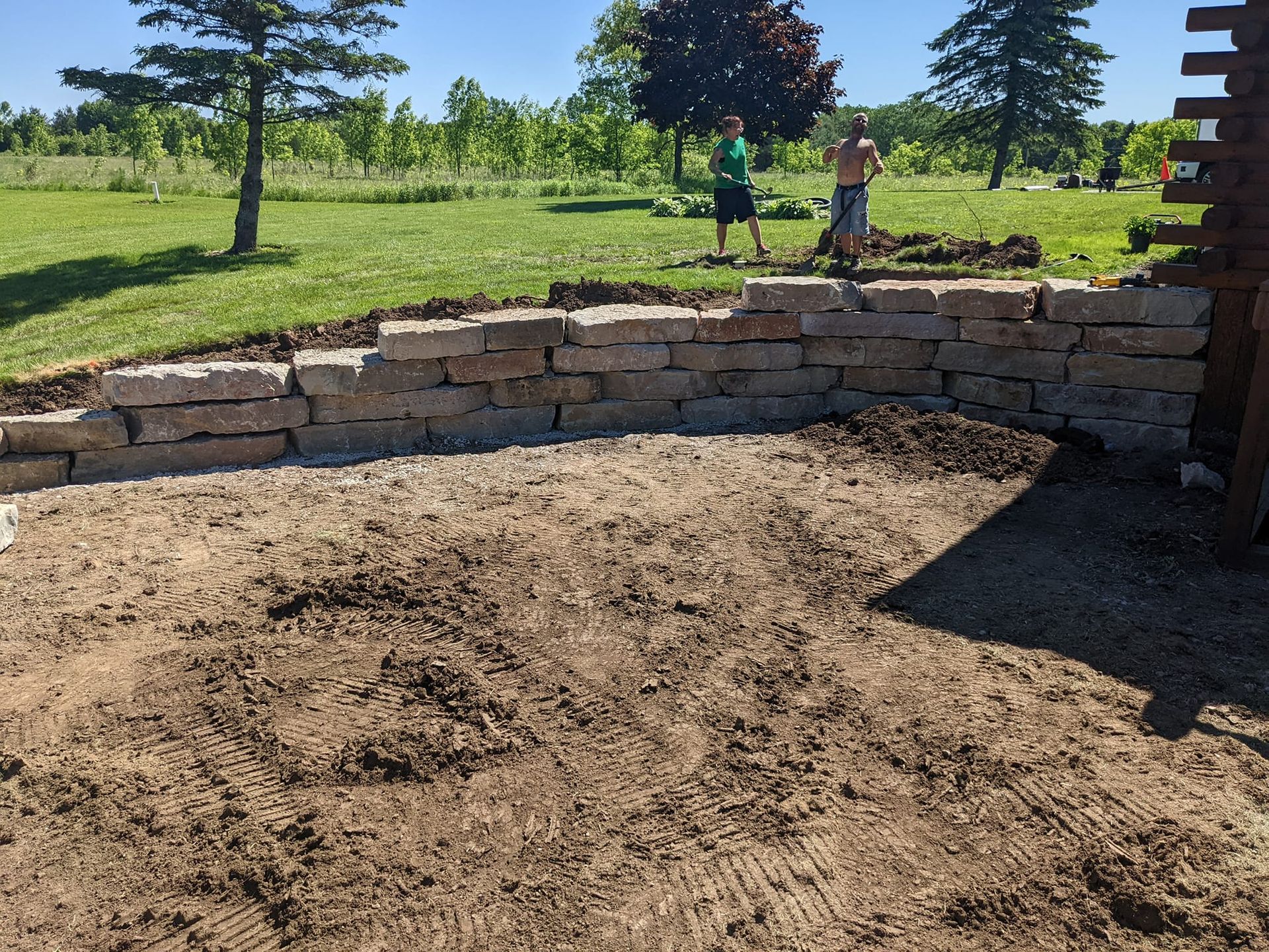 Stone retaining wall under construction in a yard; two people working in the background.