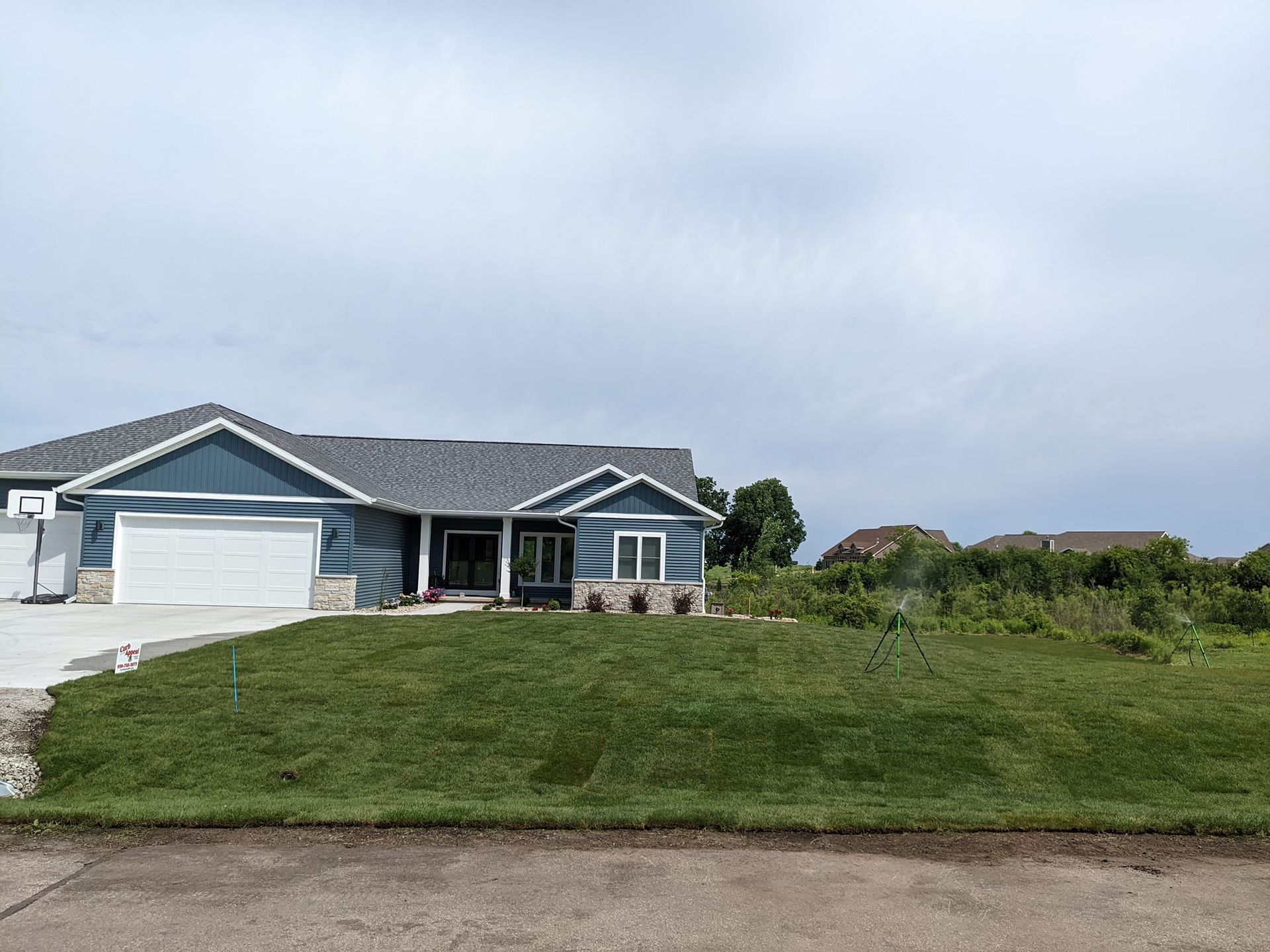 Blue house with white garage and grey roof on green lawn under cloudy sky.