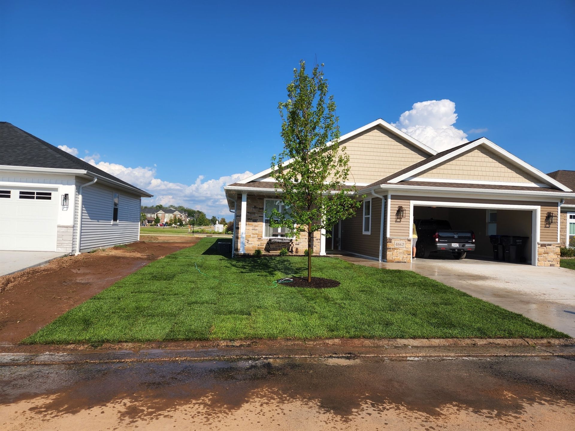 Newly sodded front yard with a young tree in front of a tan house with an open garage.