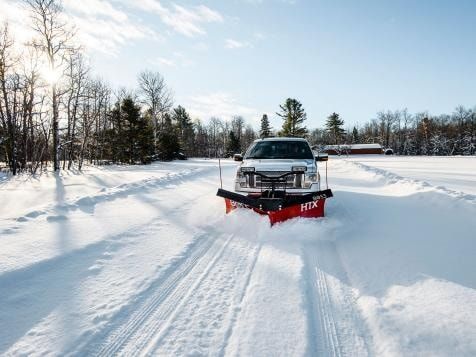 A silver pickup truck with a red snowplow clears a snow-covered road on a sunny day.