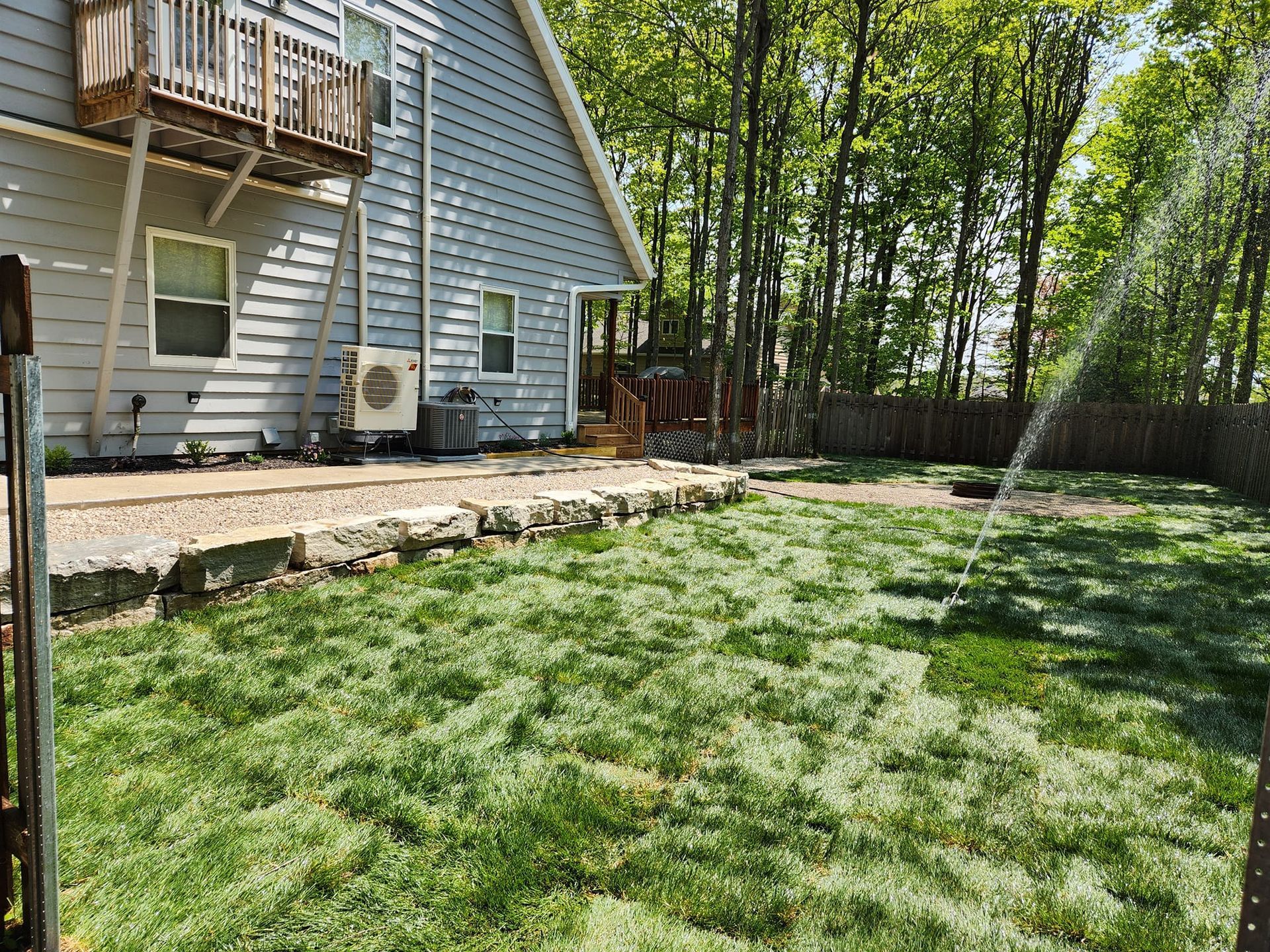 Backyard with house and sprinkler watering the green grass. Trees in the background.