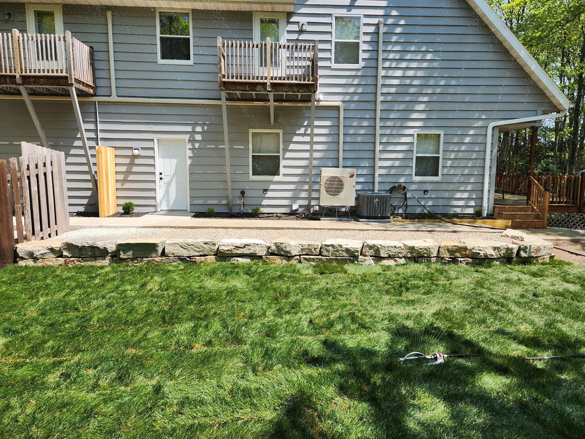 Backyard patio with stone retaining wall, grass, and a gray house with two balconies.