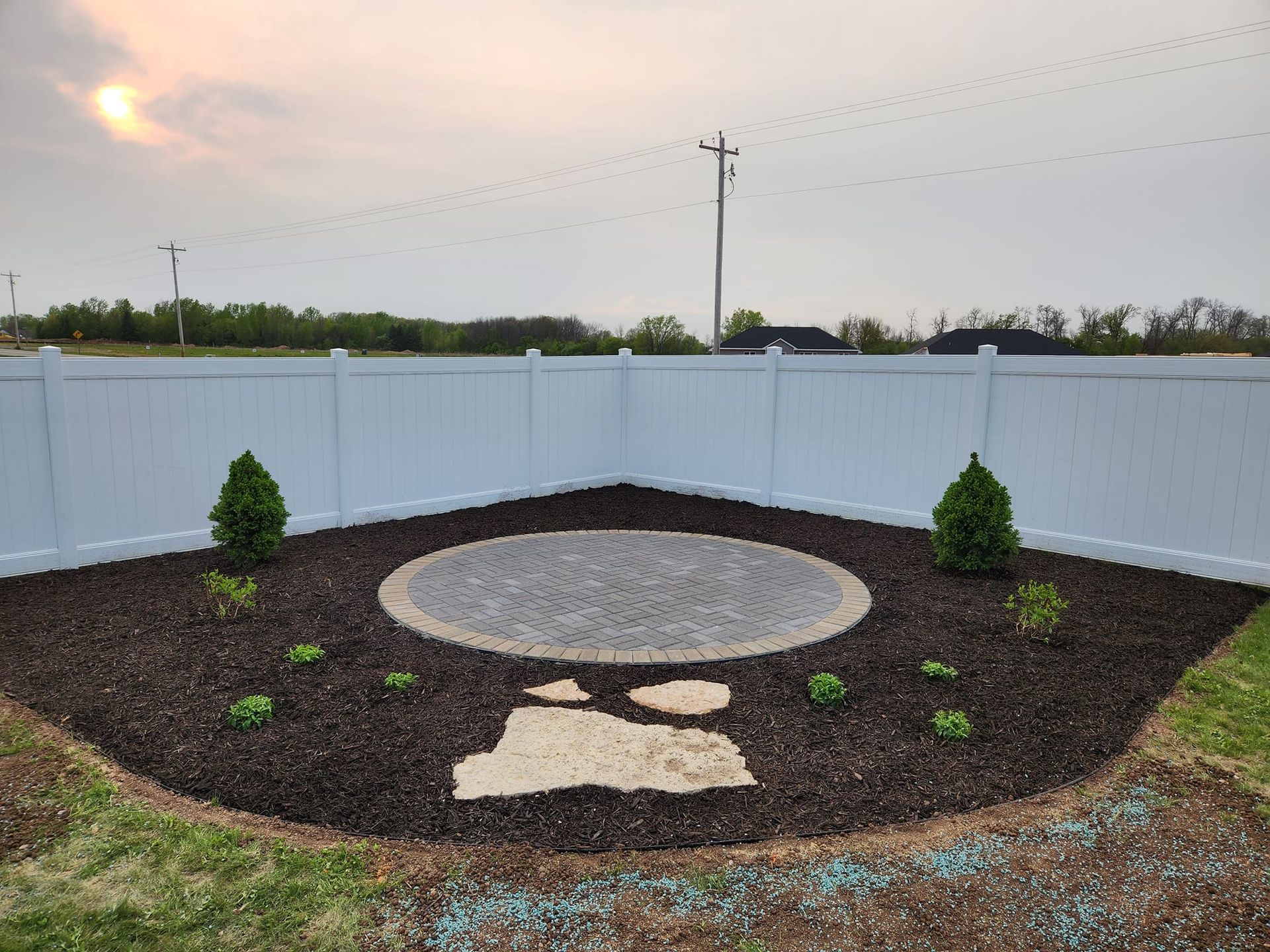 A backyard patio with a gravel seating area surrounded by mulch, plants, and a white fence.