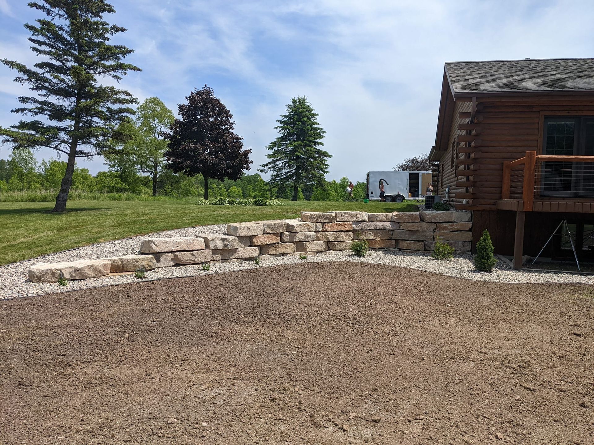 A log cabin with a stone retaining wall and gravel area; trees and sky are in the background.