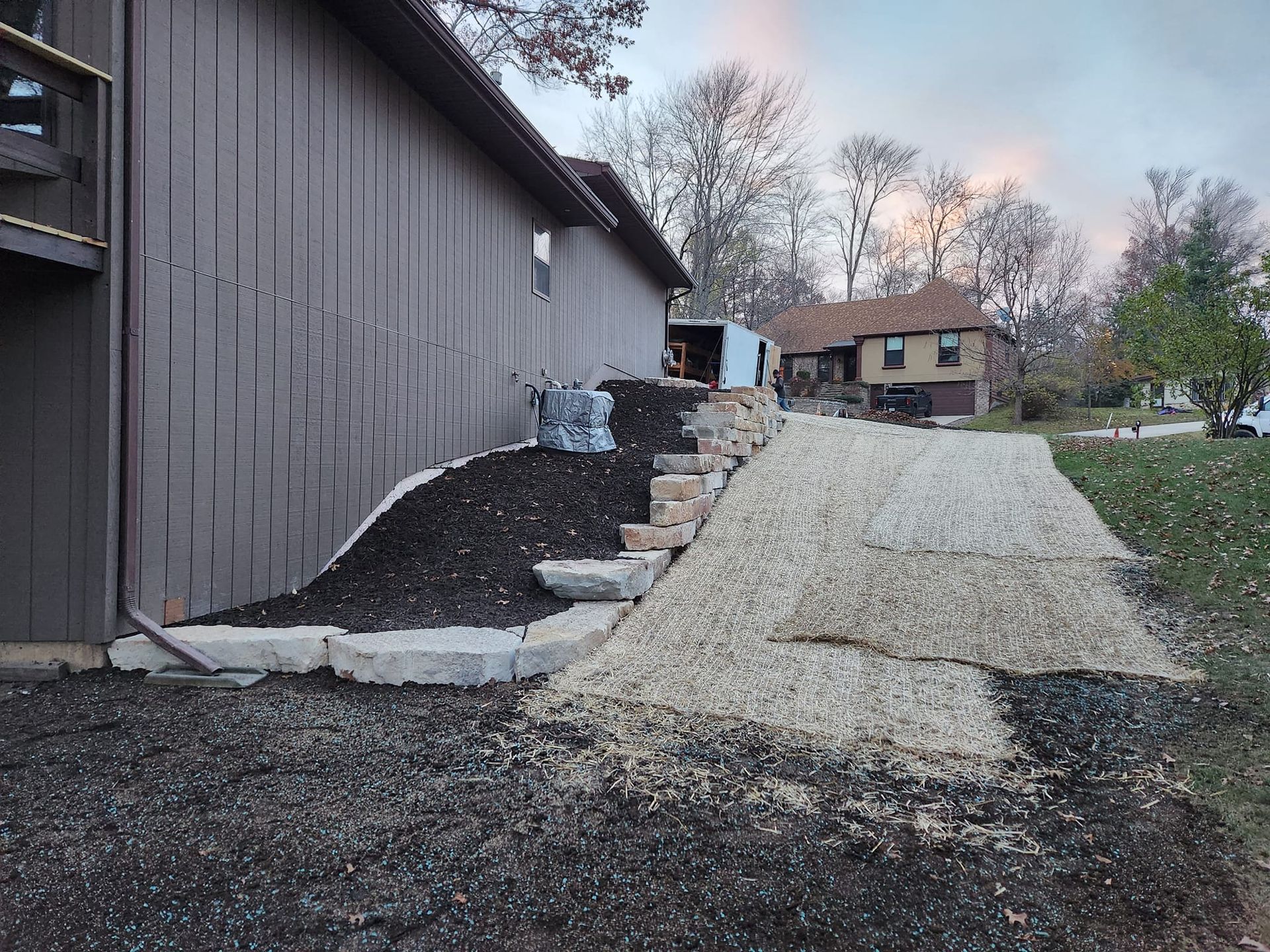 Side of a house with a stone retaining wall, steps, and gravel pathway leading up. Dark mulch and a house in the background.