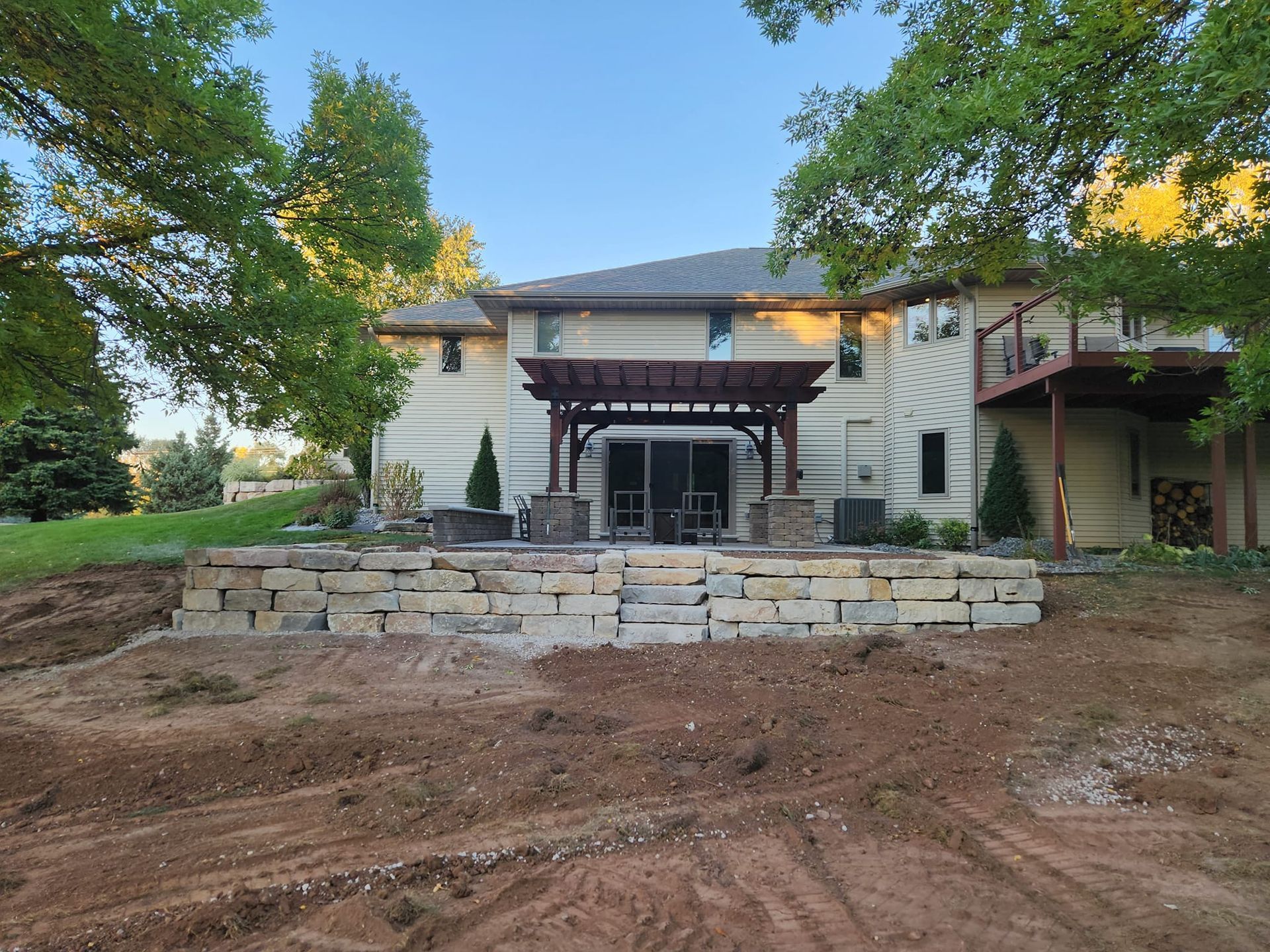 Two-story beige house with a stone patio, pergola, and retaining wall in a yard with dirt.