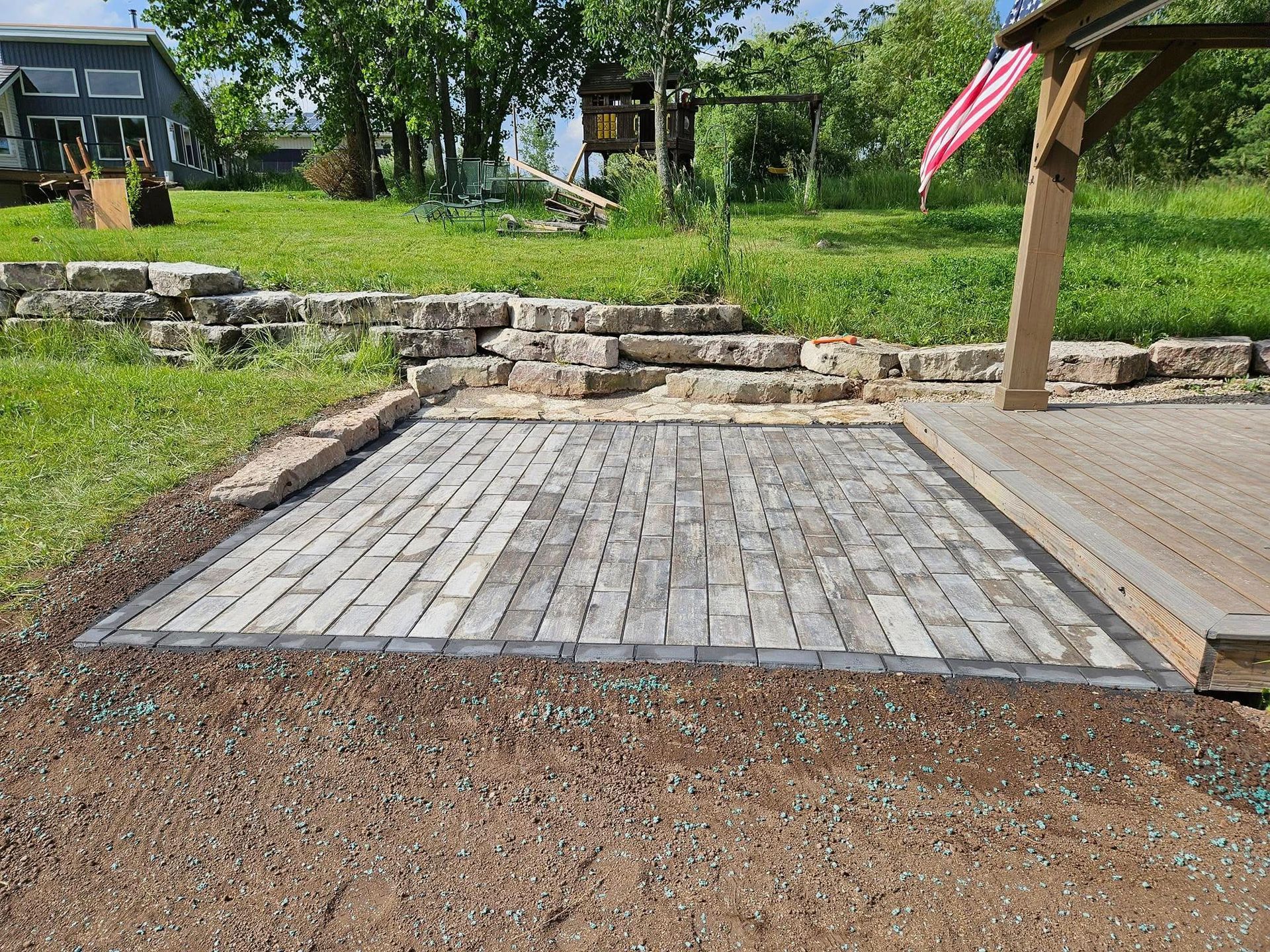 A brick patio bordered by gravel and stone with a deck and landscaping in the background.