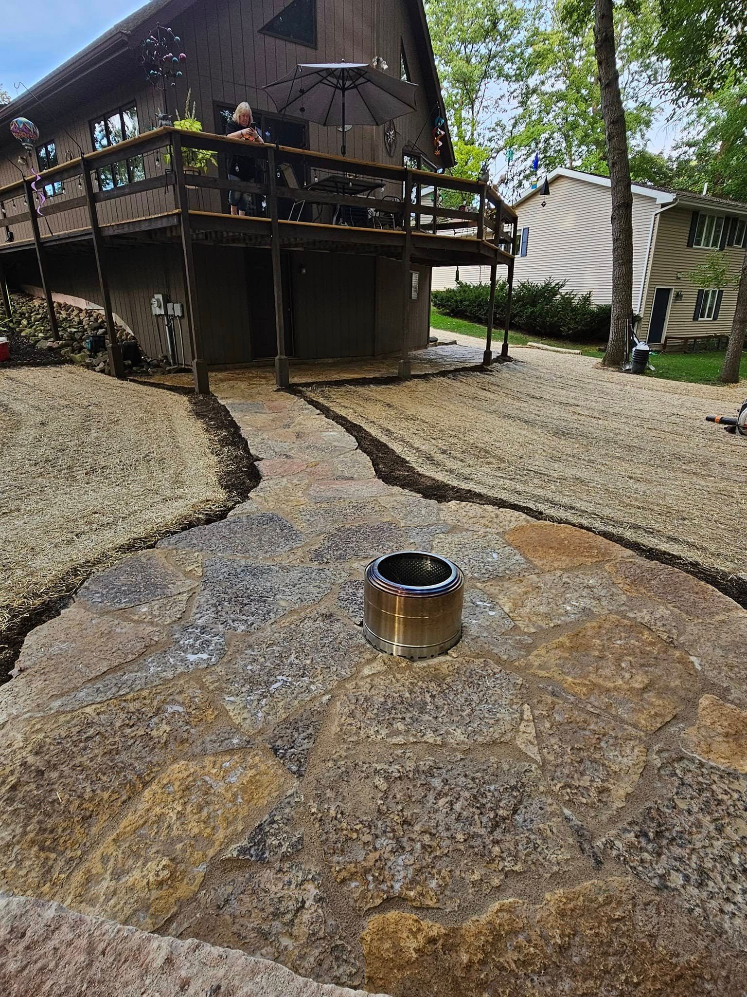 A rock patio leads to a deck. A stainless steel cylinder is centered. The surrounding area has tan gravel and a house in the background.