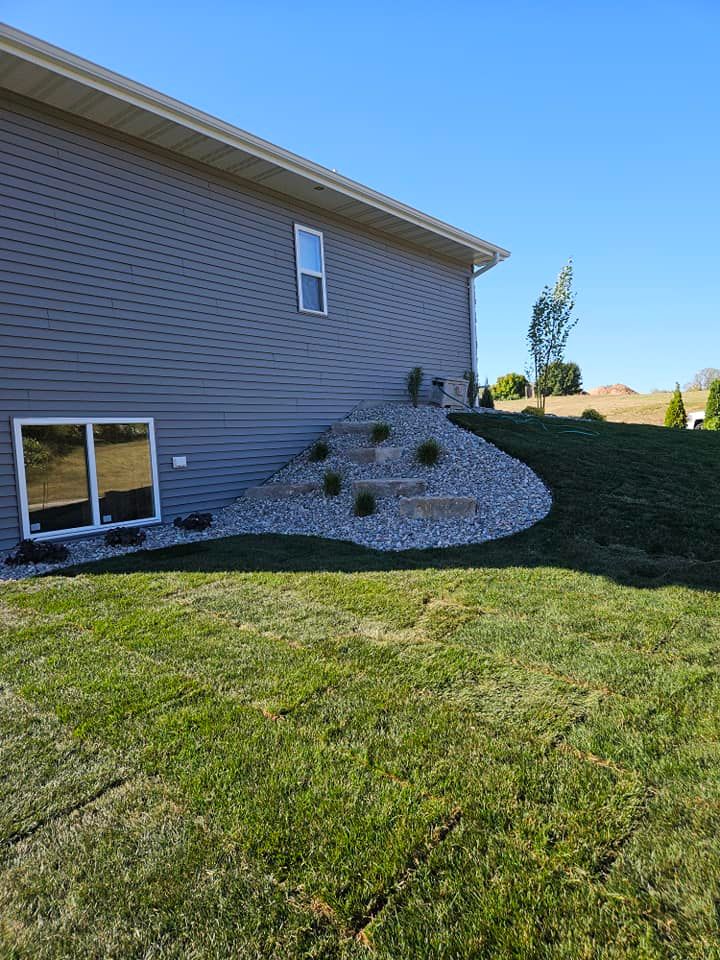Side of a house with gray siding, landscaping with rocks, steps, and small plants on a small hillside. Green grass in yard.