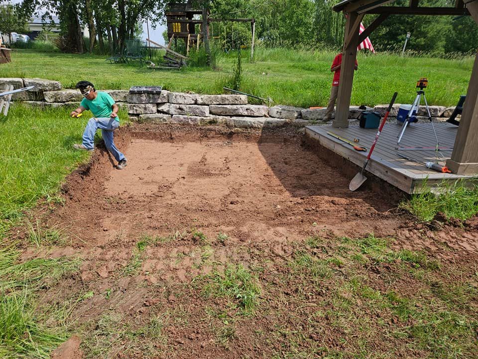 Person digging in a dirt area near a wooden gazebo, surrounded by grass and a low stone wall.