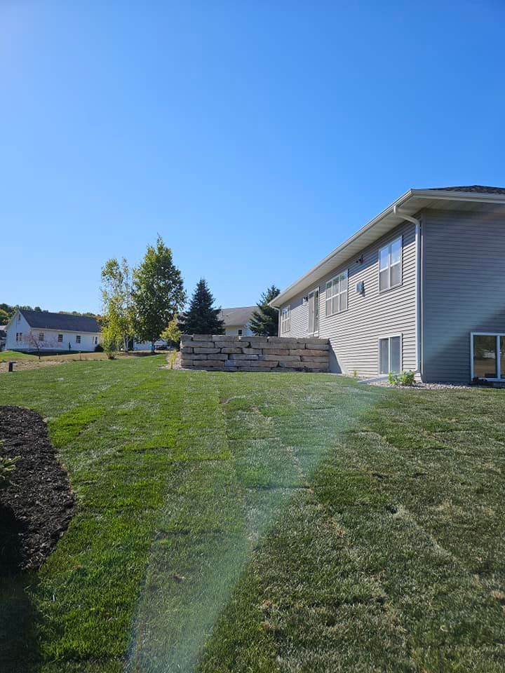 Green lawn with a two-story gray house to the right. A retaining wall in the mid-ground. Clear, blue sky.