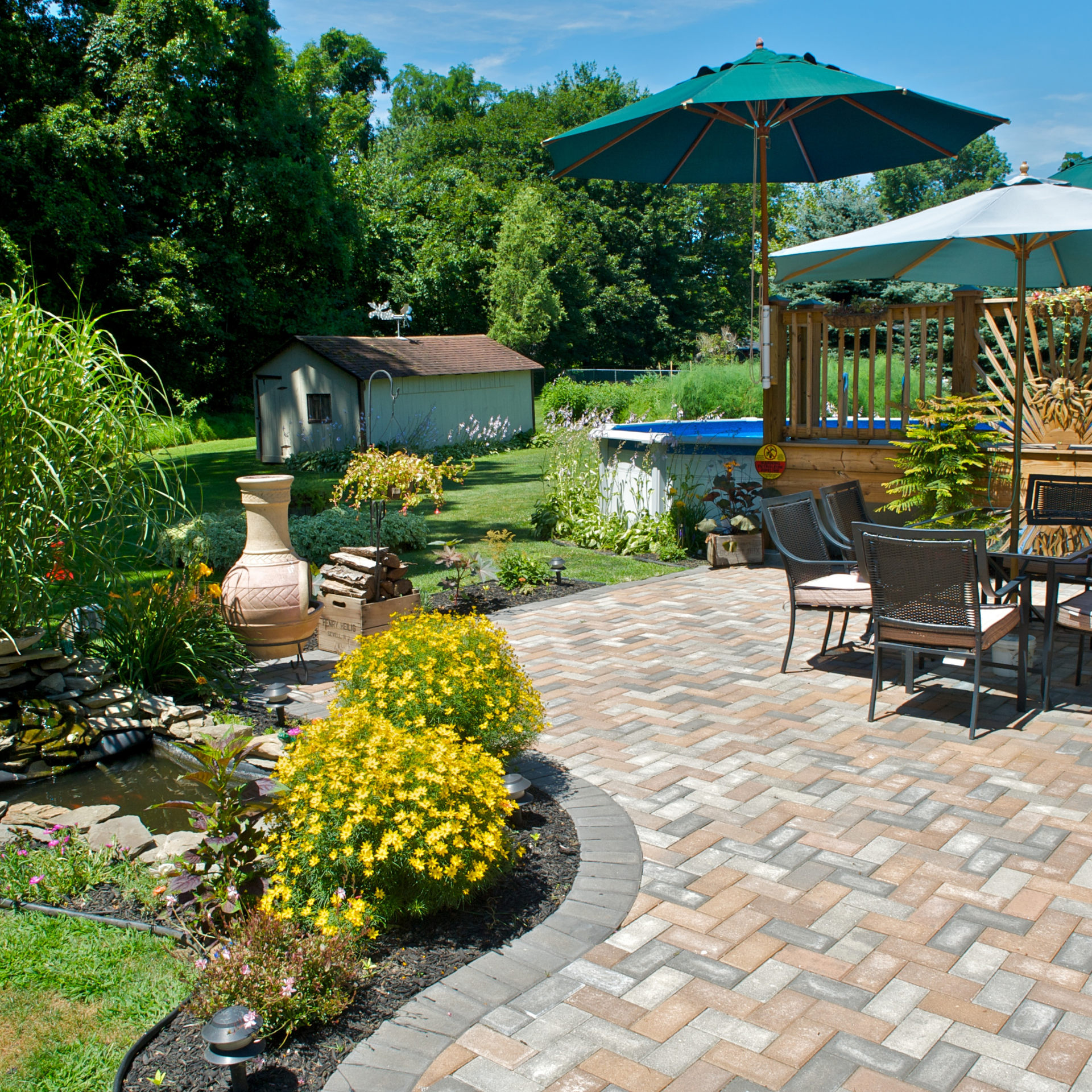 Brick patio with seating, umbrellas, and small pond in a lush, sunny backyard.