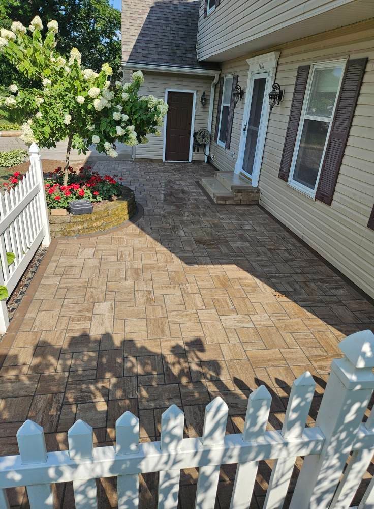 Brick patio bordered by a white picket fence, garden, and a beige house with brown trim.