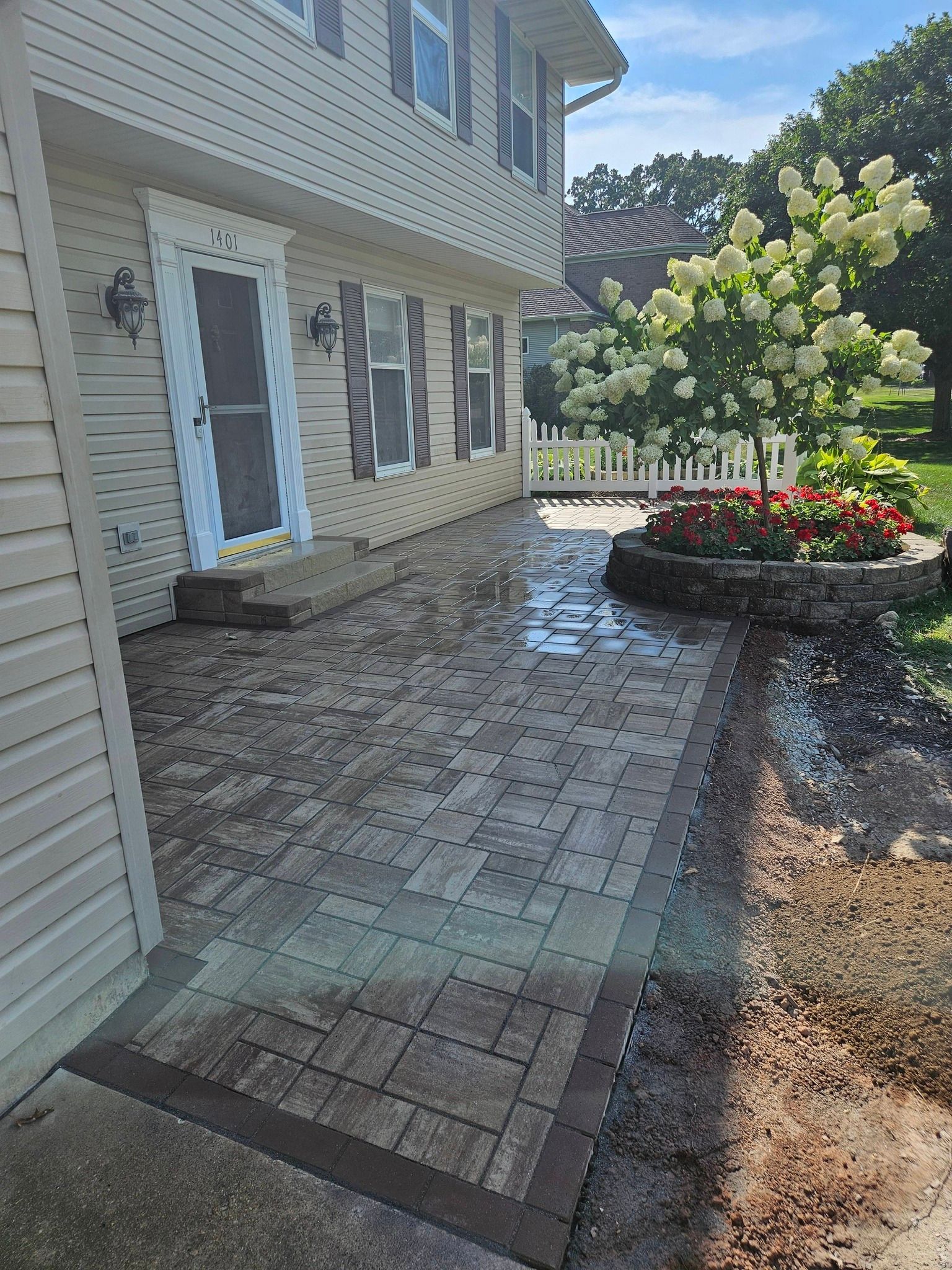 Brick patio in front of a house, with flowers and shrubs on the side.