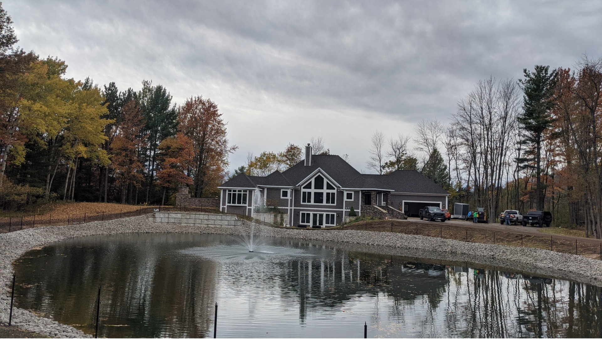 House on a pond with a fountain, surrounded by trees. Cloudy sky above.