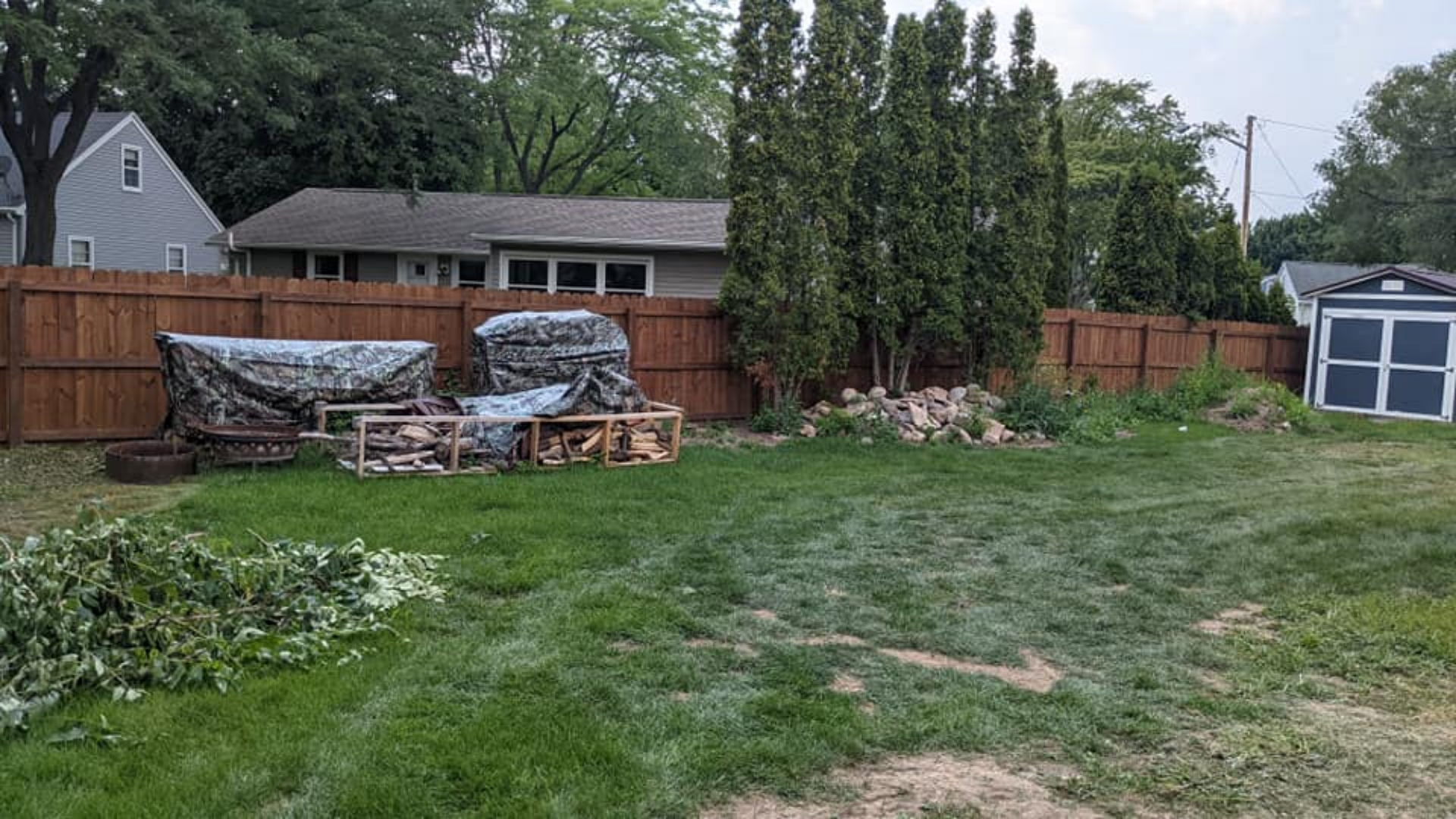Backyard with wood fence, storage, firewood, green grass, and trees.