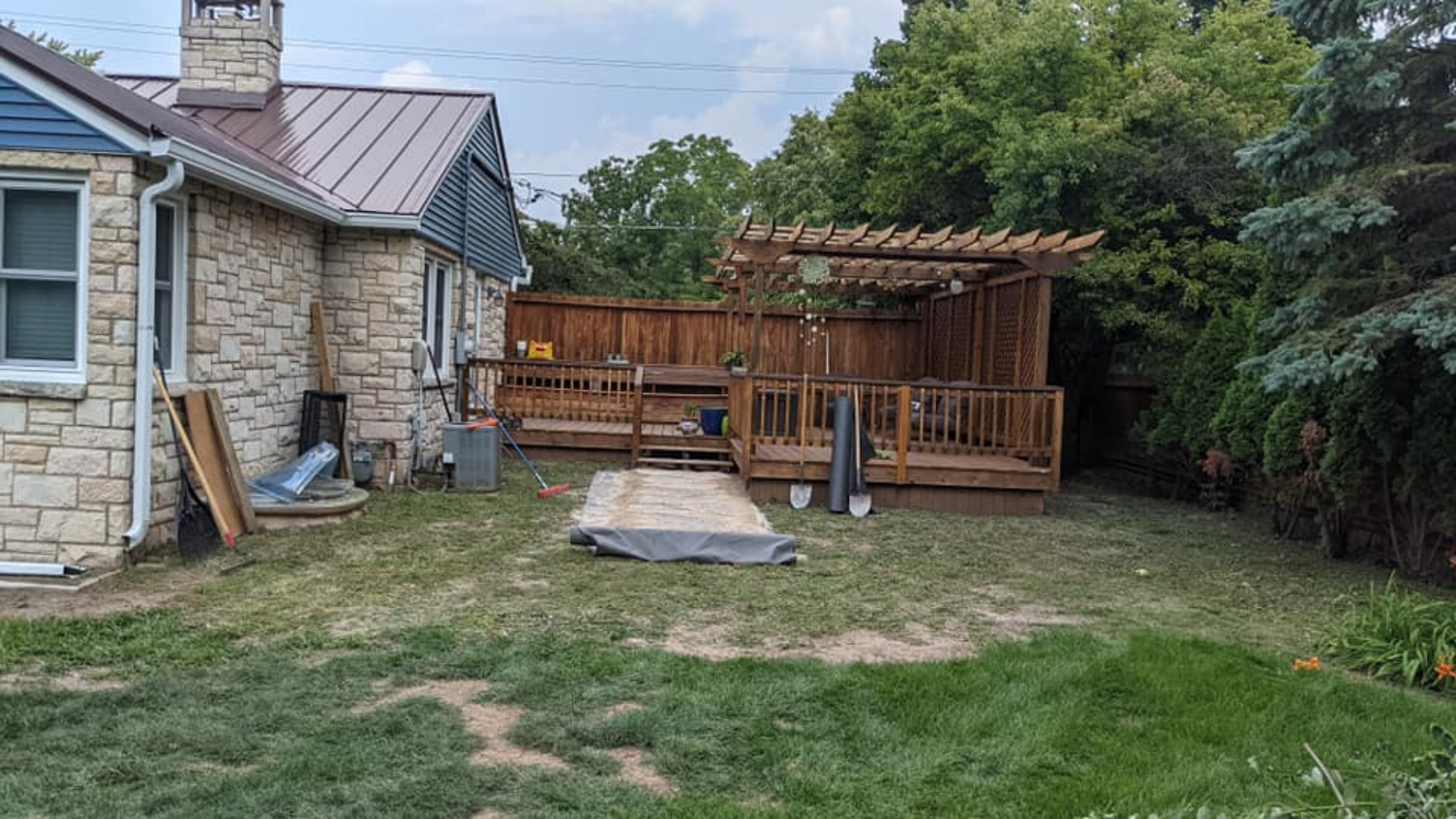 Backyard with a wooden deck, pergola, and brown fence next to a stone house and green grass.