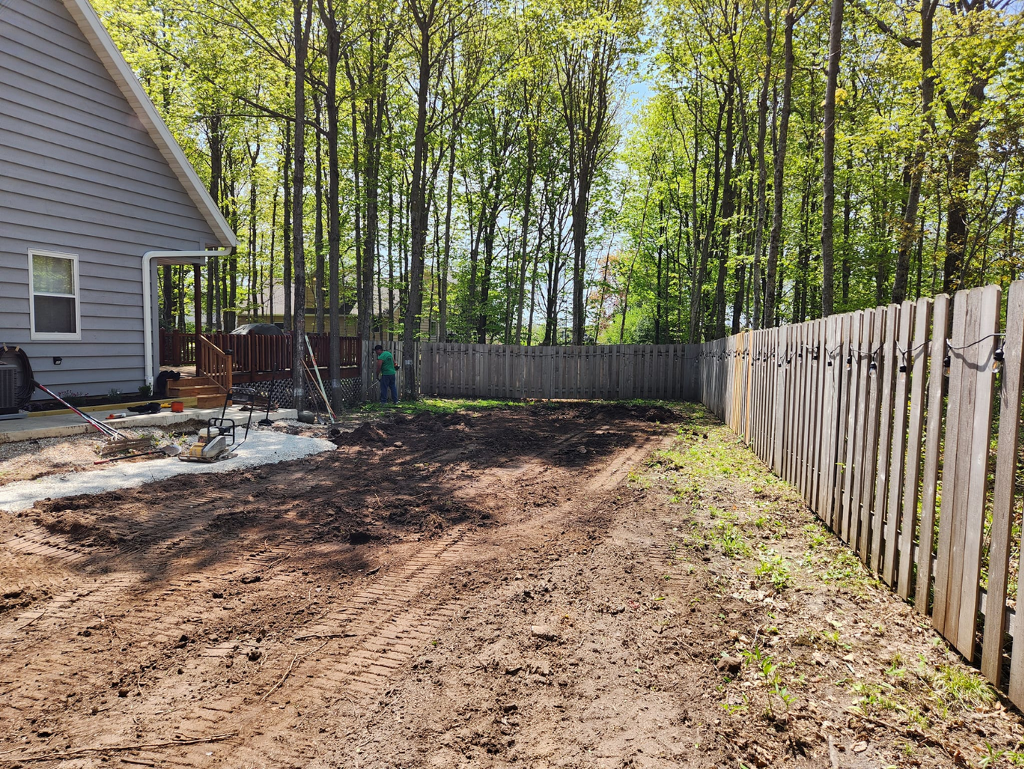 Muddy backyard cleared of vegetation, next to a wooden fence and house. Trees in the background.