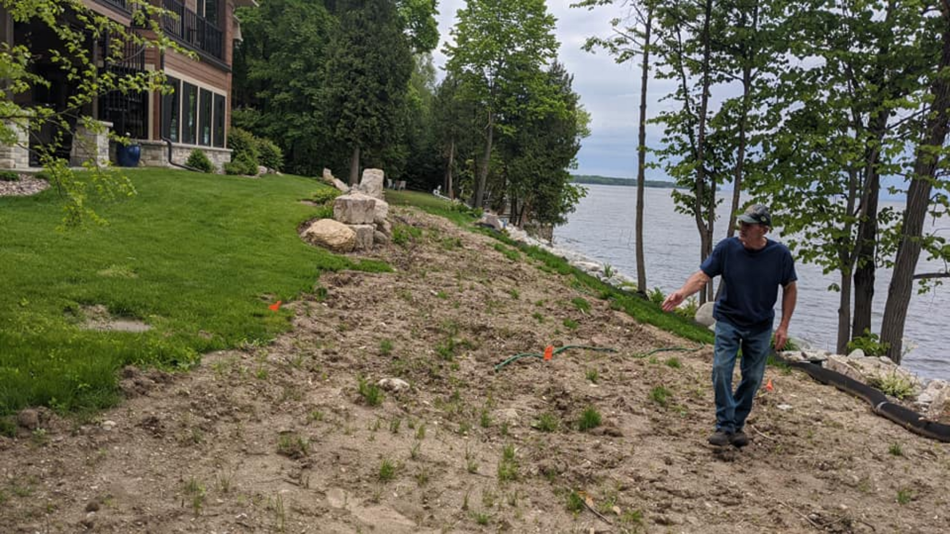 Man walks on a dirt slope near a lake, pointing. A house and trees are in the background on a cloudy day.