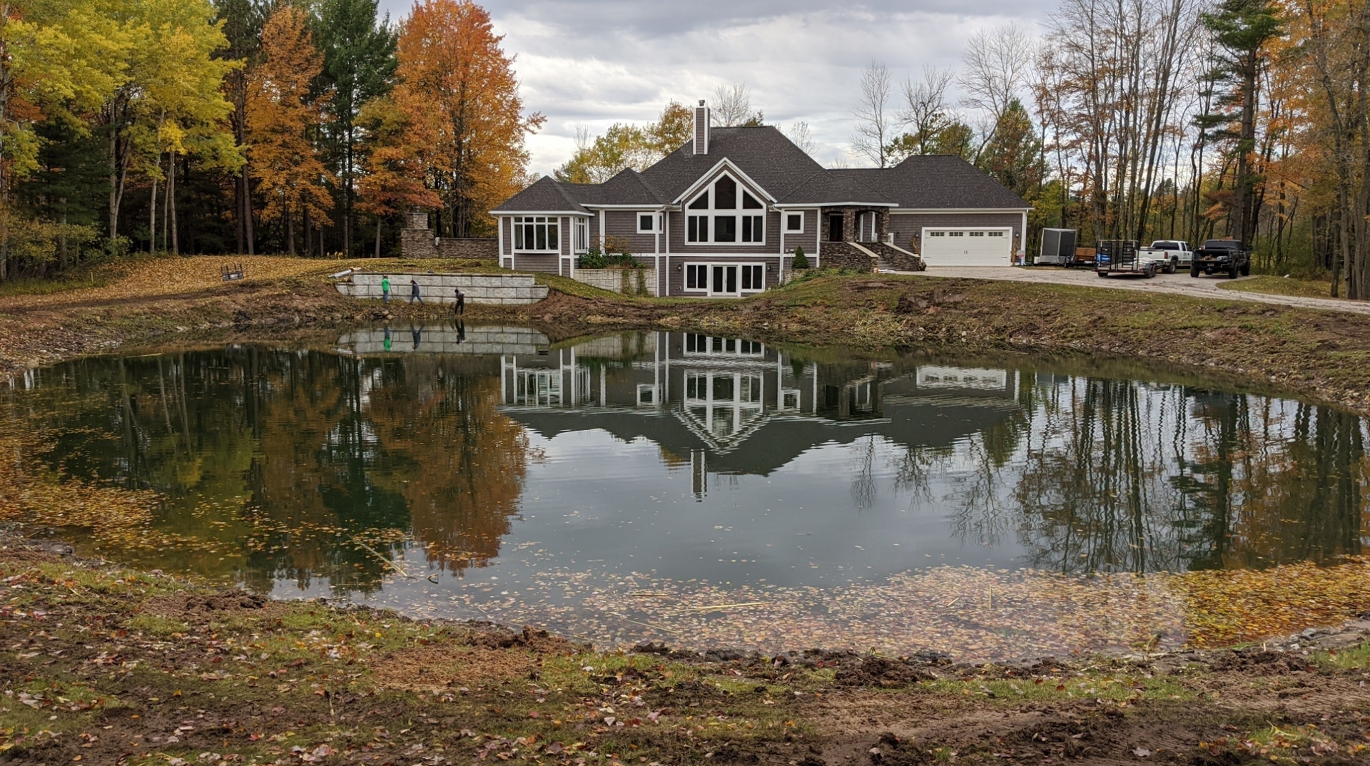 Large house reflected in a pond surrounded by fall foliage. Gray sky, brown and yellow trees, muddy shoreline.