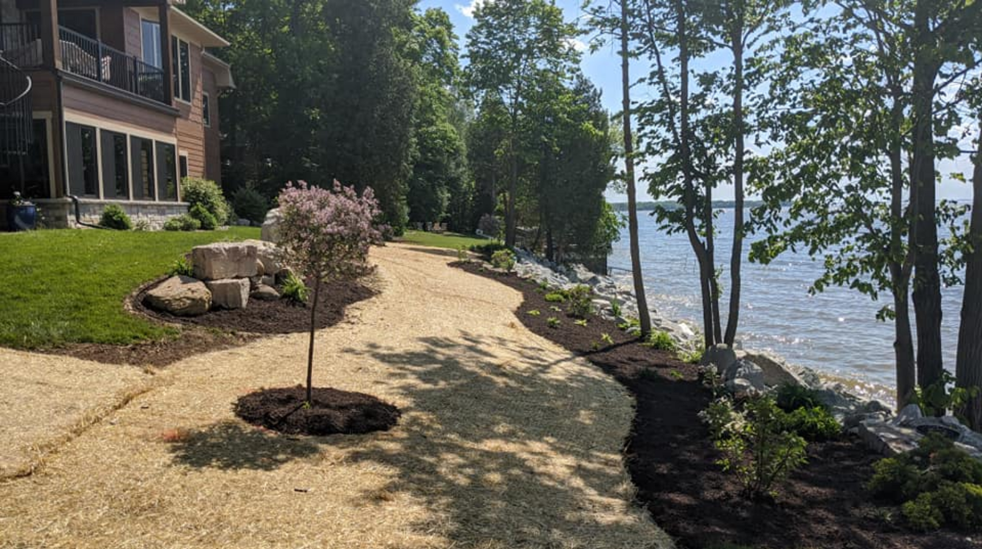 A shoreline path lined with trees and landscaping, adjacent to a large house.