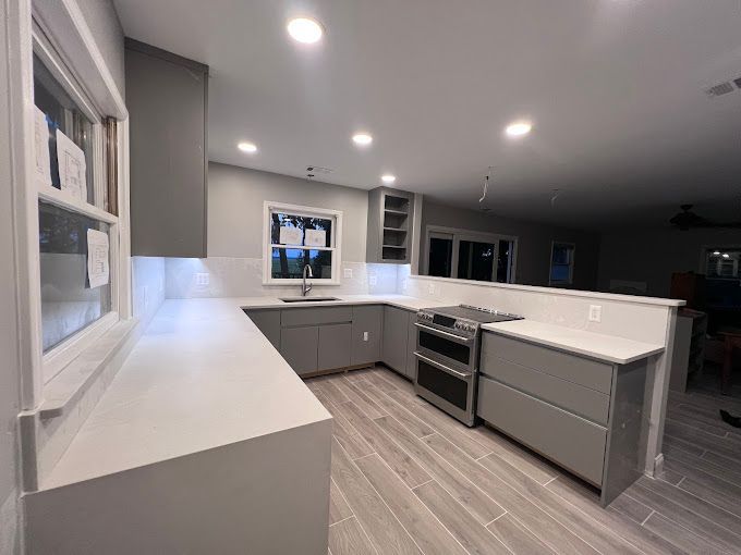 A kitchen with gray cabinets , white counter tops , a stove , and a window.