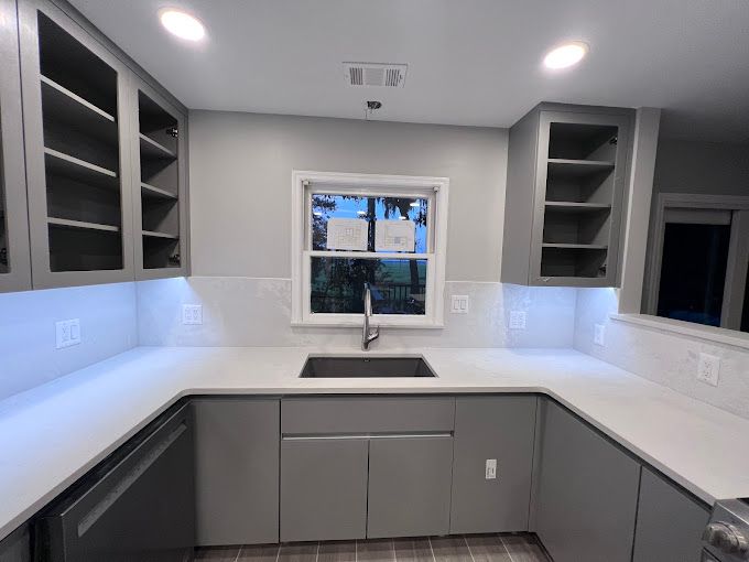 A kitchen with gray cabinets , white counter tops , a sink and a window.