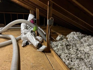 Worker in white protective suit insulating an attic with blowing hoses and loose insulation.