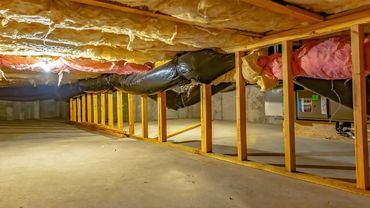 Crawlspace under a house with exposed insulation, yellow support posts, ductwork, and a concrete floor.
